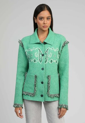 Jeune femme portant une veste verte boutonnée avec des franges et des motifs brodés blancs, associée à un pantalon gris clair, en position neutre.