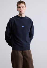 Μπλε σκούρο πουλόβερ με ριμπ κολάρο και τελείωμα, με μικρό μπλ ε ορθογώνιο λογότυπο. Συνδυασμένο με καφέ παντελόνι. Ομαλή υφή.