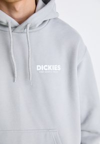 Ljusgrå hoodie med dragsnören, har en framficka och vit text: "DICKIES, FORT WORTH, TEXAS" tryckt på bröstet.
