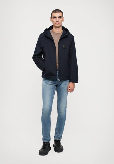 Navy hooded jacket with a front zipper, textured brown shirt beneath, light blue jeans, and black lace-up shoes. Minimalistic design.