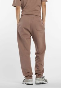 Pantalones de chándal marrones con cinturilla elástica y tobillos ajustados, que presentan un bolsillo trasero. Combinados con zapatillas grises. Textura de tela suave.