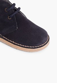 Zapato de ante azul marino con punta redondeada, suela de goma marrón y costuras en contraste. Incluye dos cordones oscuros a través de las ojales.