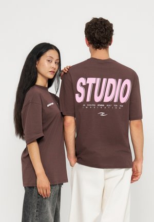En kvinde og en mand har matchende brune T-shirts på; på mandens ryg er stor lyserød tekst med teksten "STUDIO" og mindre tekst under.