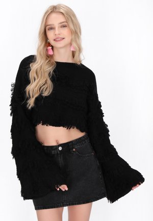 Schwarzer, kurz geschnittener Pullover mit strukturierten Rüschendetails an den Ärmeln und am Saum, kombiniert mit einem schwarzen Jeansrock. Der Stoff wirkt weich und leicht abgenutzt.