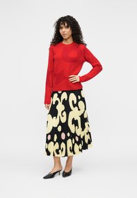 Κόκκινο πλεκτό πουλόβερ με αφηρημένα σχέδια, σε συνδυασμό με μαύρη πλισέ φούστα με κίτρινα και ροζ μοτίβα. Μαύρα παπούτσια με μυτερή μύτη.