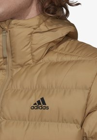 Tan pufferjakke med lynlås foran og hætte. Har rundede skuldre, quiltet tekstur og et sort Adidas-logo på brystet.