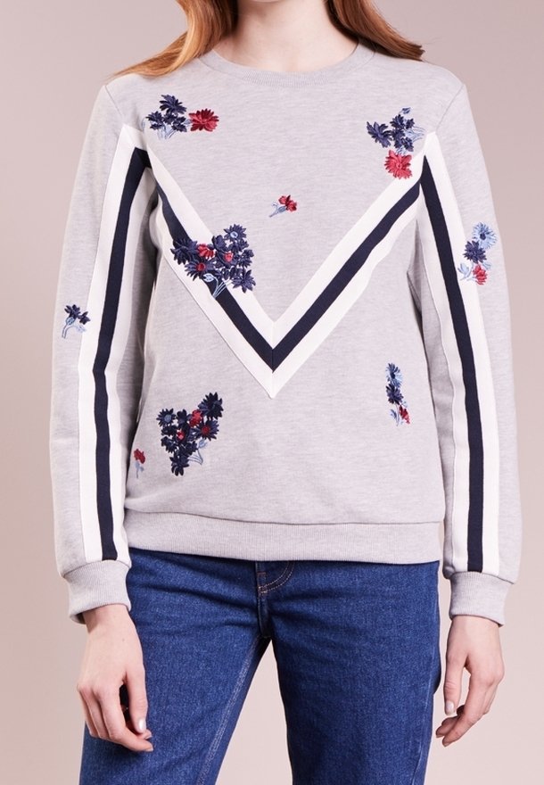 Sweat-shirt gris clair avec des bandes chevrons blanches et noires et une petite broderie florale rouge et bleue, porté avec un jean bleu.