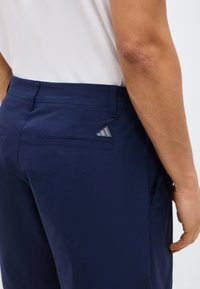 Κοντινή λήψη ενός ατόμου που φοράει σκούρο μπλε παντελόνι με ένα μικρό λογότυπο Adidas κοντά στην δεξιά πίσω τσέπη και ένα λευκό πουκάμισο.