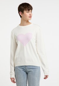 Pull en blanc avec un motif de cœur rose pastel côtelé au centre. Présente un col rond et des poignets côtelés, fabriqué dans un matériau en tricot doux.