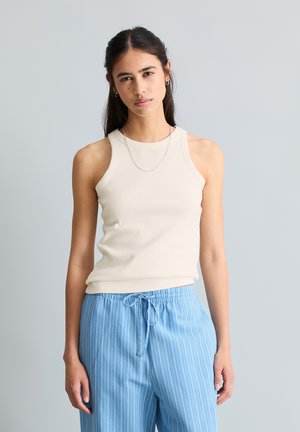 Kobieta z długimi ciemnymi włosami, ubrana w beżowy top bez rękawów i jasno-niebieskie paskowane spodnie ze ściągaczem, stojąca na tle jednolitego szarego tła.