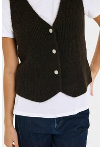 Zwarte gebreide mouwloze vest met drie bloemvormige parelknoopjes over een witte T-shirt met korte mouwen, gecombineerd met donkerblauwe spijkerbroek.