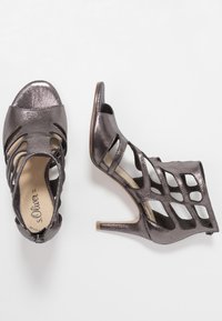 Korkease sandaletit kiilakorkoilla kiiltävästä tummasta metallimateriaalista. Varustettu leikattu muotoilu, avonainen varvas ja kapea stiletto-korko. Säädettävä nilkkahihna.