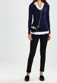 Marineblauwe v-hals trui met zilveren ritssluitingen, witte blouse eronder, zwarte skinny broek en zwarte schoudertas met bloemenpatroon.