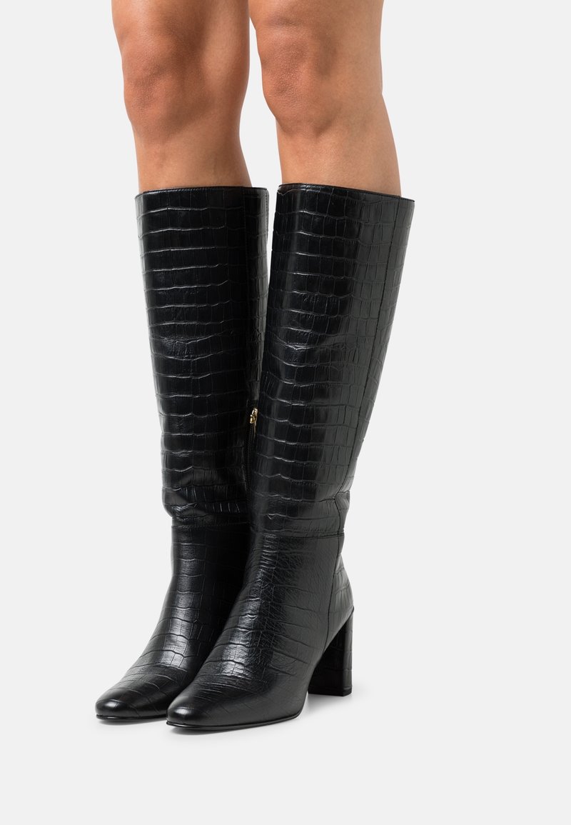 Vue des jambes portant des bottes noires en cuir à texture crocodile, montant jusqu'au genou, avec des talons carrés, sur un fond blanc.