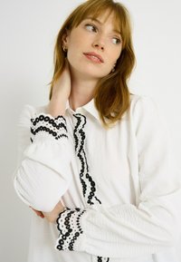 Blusa blanca con un borde de encaje negro en el cuello y en los puños. La tela tiene una textura suave con un aspecto arrugado.