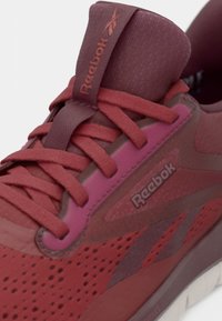 Încălțăminte sport Reebok în nuanțe de roșu și roz, prezentând un superior din mesh respirabil, accente texturate și un sistem de șireturi proeminent.