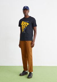 T-shirt de algodão preto com o logótipo amarelo "JUST DO IT", calças rectas castanhas e sapatos pretos com meias brancas. O boné combina com a t-shirt.
