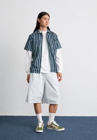 Un jeune homme se tenant sur un tapis bleu, portant une chemise à rayures à manches courtes par-dessus une chemise à manches longues blanche, un short blanc et des baskets vertes à motifs.