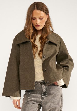 Femme aux longs cheveux ondulés portant une veste marron oversize boutonnée sur un pull crème et un jean gris délavé, debout avec une main dans la poche.