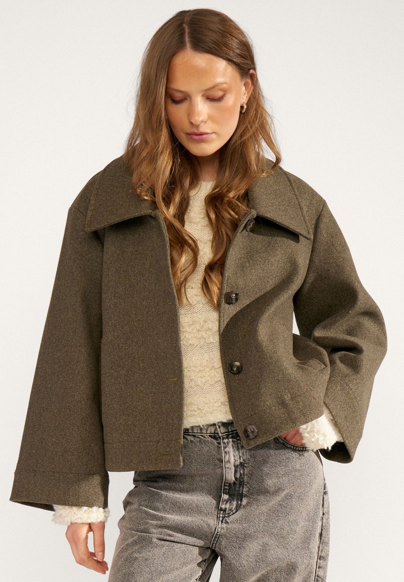 Femme aux longs cheveux ondulés portant une veste marron oversize boutonnée sur un pull crème et un jean gris délavé, debout avec une main dans la poche.
