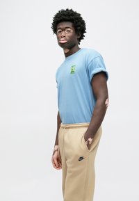 Világoskék pamut póló felhajtott ujjakkal, a mellkason zöld logóval. Bézs színű melegítőnadrág fekete Nike logóval. Sima textúra.