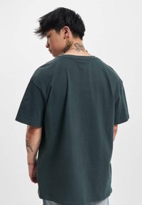 Camiseta de algodón verde oscuro con mangas cortas, un corte relajado y cuello redondo. La parte trasera presenta una textura suave, sin patrones y con un diseño sencillo.
