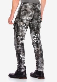 Schwarze und graue Cargo-Hosen mit Graffiti-ähnlichem weißen Text. Verfügen über Taschen, eine strukturierte Oberfläche und einen Slim-Fit, kombiniert mit schwarzen Stiefeln.