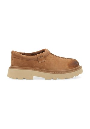 Zapato marrón de ante tipo slip-on con un upper texturizado, suela de goma clara en contraste y costuras visibles a lo largo del borde. Etiquetado como "UGG".