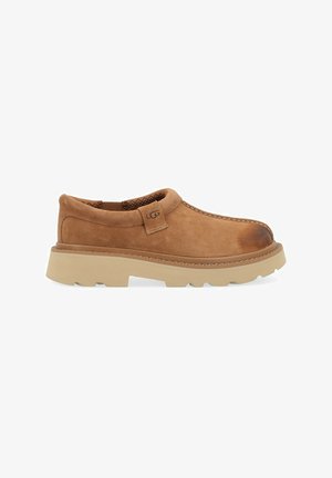 Scarpa slip-on in suede marrone con tomaia texturizzata, suola in gomma chiara a contrasto e cuciture visibili lungo il bordo. Etichettata "UGG".