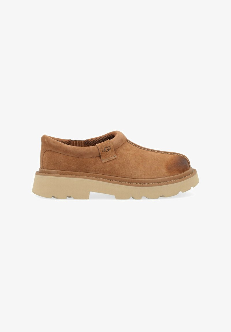 Scarpa slip-on in suede marrone con tomaia texturizzata, suola in gomma chiara a contrasto e cuciture visibili lungo il bordo. Etichettata "UGG".