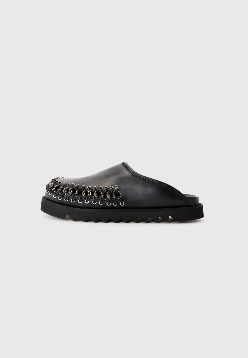 Chaussure noire en cuir à enfiler avec des détails de couture en œillets argentés et une semelle noire dentelée, présentée en profil latéral sur fond blanc.