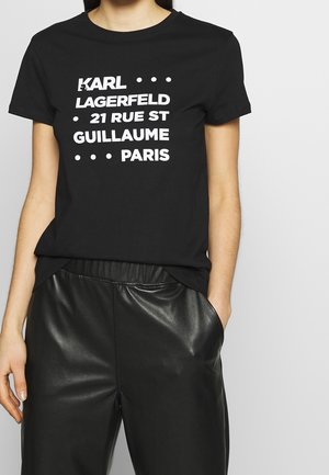 Žena nosící černé tričko s bílým nápisem "KARL LAGERFELD 21 RUE ST GUILLAUME PARIS" a černé kožené kalhoty.