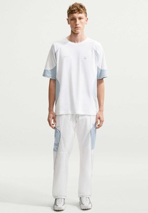 Jeune homme debout face à l'objectif, portant un t-shirt blanc et un pantalon blanc avec des empiècements bleu pâle, et des baskets blanches sur un fond uni.