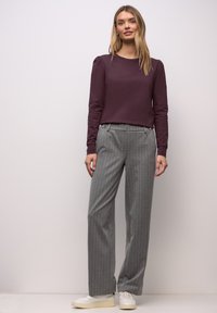 Frau steht und trägt ein dunkelviolettes Langarmshirt, graue, breit geschnittene Hosen mit Nadelstreifen und weiße Turnschuhe vor einem einfarbigen Hintergrund.