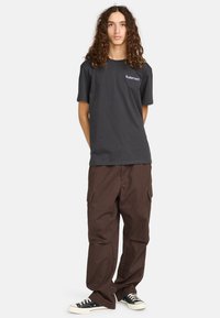 Schwarzes Baumwoll-T-Shirt mit einem Logo auf der linken Brust, kombiniert mit braunen Cargo-Hosen mit mehreren Taschen und einem lässigen Schnitt, getragen mit schwarzen Sneakers.
