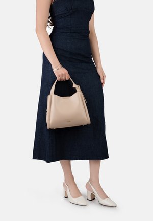 Femme en robe sombre sans manches tenant un sac à main beige, portant des chaussures à talon bloc blanches à bride arrière sur fond blanc.