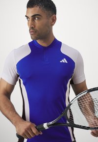 Blauw sportshirt met korte mouwen, witte accenten en een halve rits. Het model houdt een tennisracket vast met een zwarte handgreep.