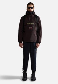 Brown waterproof jacket with a front pocket, logo, and Norwegian flag patch, paired with navy pants and black sneakers. Sunglasses worn.