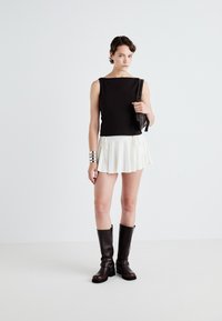 Top senza maniche nero, gonna plissettata bianca, bracciale in argento e stivali al ginocchio marroni. Modella tiene in mano una borsa nera.