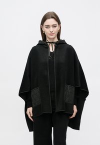 Mustast pehmest kangast kapuutsiga viig, millel on tikitud küljetaskud, lukuga sulgemine ja dekoratiivne lips kaeluse juures.