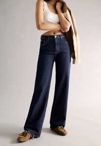 Wide-leg dark denim jeans with a high waist, featuring front pockets and a button closure. Paired with a white tank top and beige jacket.