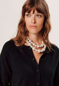 Schwarzer Cardigan mit runden Knöpfen, der drei Lagen großer, glänzender, weißer Perlen am Hals präsentiert, die ihre glatte Textur betonen.