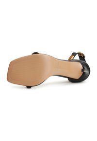 Sandalia de tacón negro con parte superior de cuero suave, correa ajustable y detalle de hebilla dorada. Suela plana beige con ligera textura.
