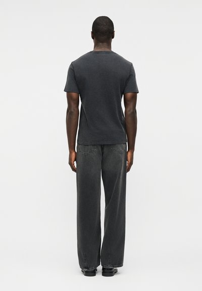 Άνδρας που στέκεται με την πλάτη προς την κάμερα, φοράει σκούρο γκρι κοντομάνικο μπλουζάκι, μαύρο χαλαρό τζιν και μαύρα παπούτσια σε απλό ανοιχτόχρωμο φόντο.