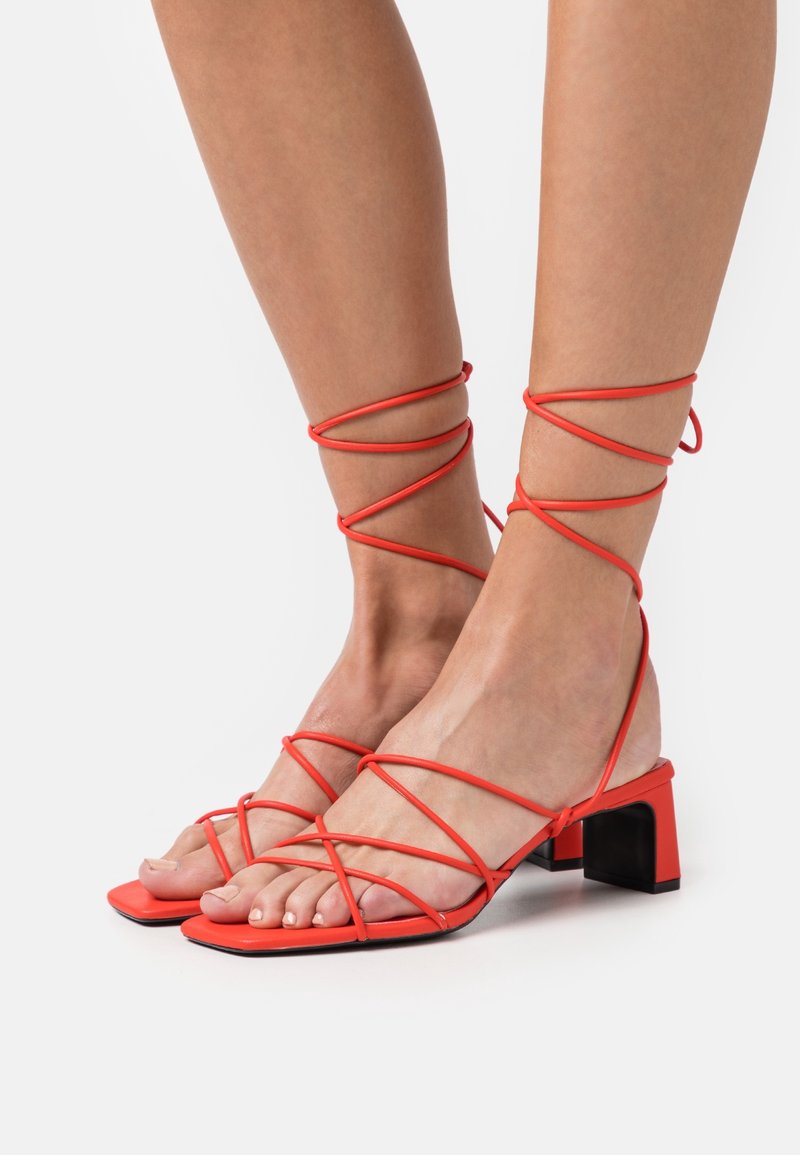 Sandales rouges à lanières avec un talon bloc et une semelle plate. Les lanières s'enroulent autour de la cheville, avec une texture lisse et un design à bout carré.