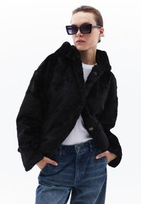 Zwarte nepbontjas met een kraag en knopen aan de voorkant, gecombineerd met een wit t-shirt en denim jeans. Grote zwarte zonnebril maakt de look compleet.