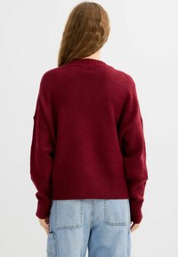 Suéter de punto burdeos con un ajuste relajado, puños y dobladillo acanalados, y una textura suave. Se lleva con pantalones cargo azul claro.