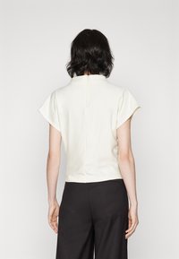 Top crema a maniche corte con collo alto, texture liscia e dettagli di cucitura sul retro, abbinato a pantaloni neri a gamba larga.
