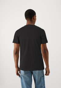 Tricou negru din bumbac, cu guler rotund, mâneci scurte și un detaliu cu cusătură centrală pe spate. Este purtat cu pantaloni cargo de culoare albastru deschis.