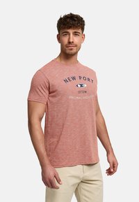 Homme portant un T-shirt rose à manches courtes avec le texte "New Port", pantalon beige, debout devant un fond clair uni.